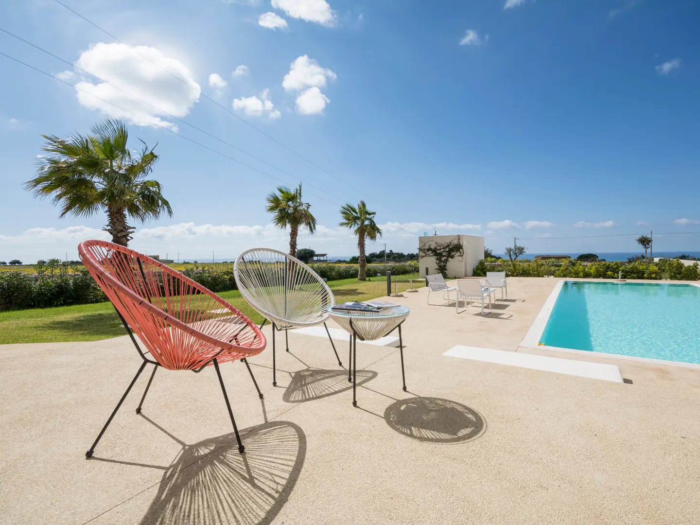Outdoor patio with a pool. Two colorful chairs and a small table sit on the patio. Palm trees and a blue sky are in the background.