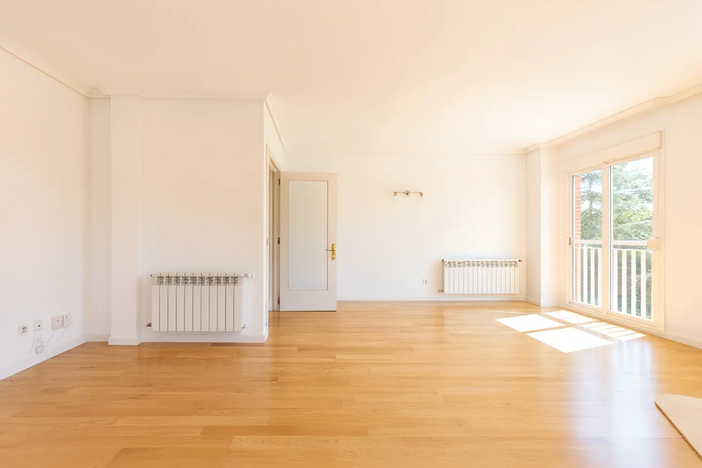 Bright, empty room with light wood floors, white walls, and a door. A window with a balcony lets in sunlight. Radiators are mounted on the walls.
