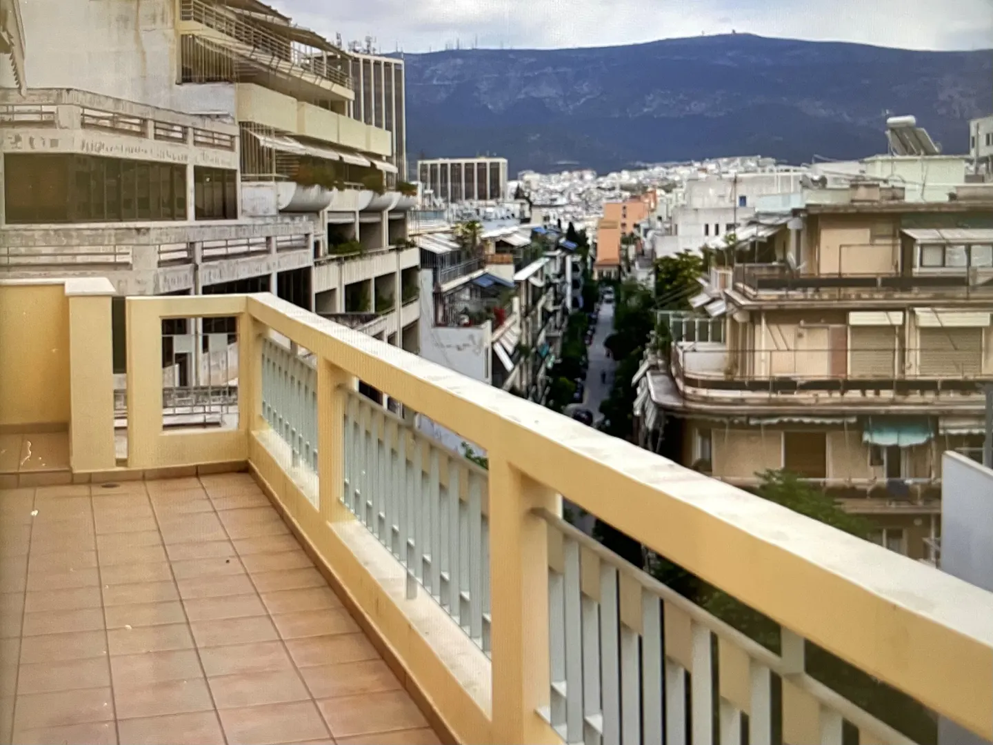 View from a tiled balcony with a beige railing overlooking a city street with buildings and mountains in the background.