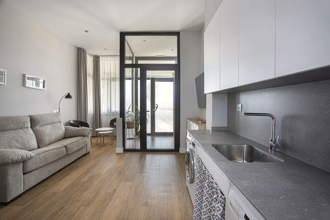 Bright, modern apartment interior with a gray sofa, wood floors, and a kitchen area with white cabinets and a gray countertop. A black-framed glass door leads to a balcony.