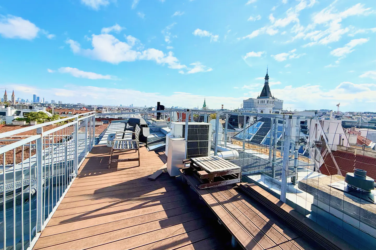 Rooftop view with wooden deck, metal railings, and city skyline under a blue sky with scattered clouds. Air conditioning units and seating are visible.