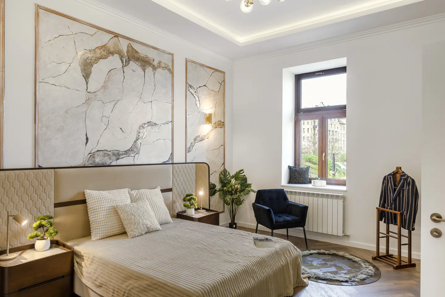 A bright bedroom with a beige bed, white walls, and large abstract art. A blue chair sits by the window, with a striped jacket on a stand nearby.