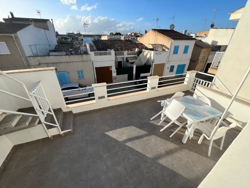 Rooftop patio with a white table and chairs, gray tile floor, and white railing. Houses with blue shutters are in the background.