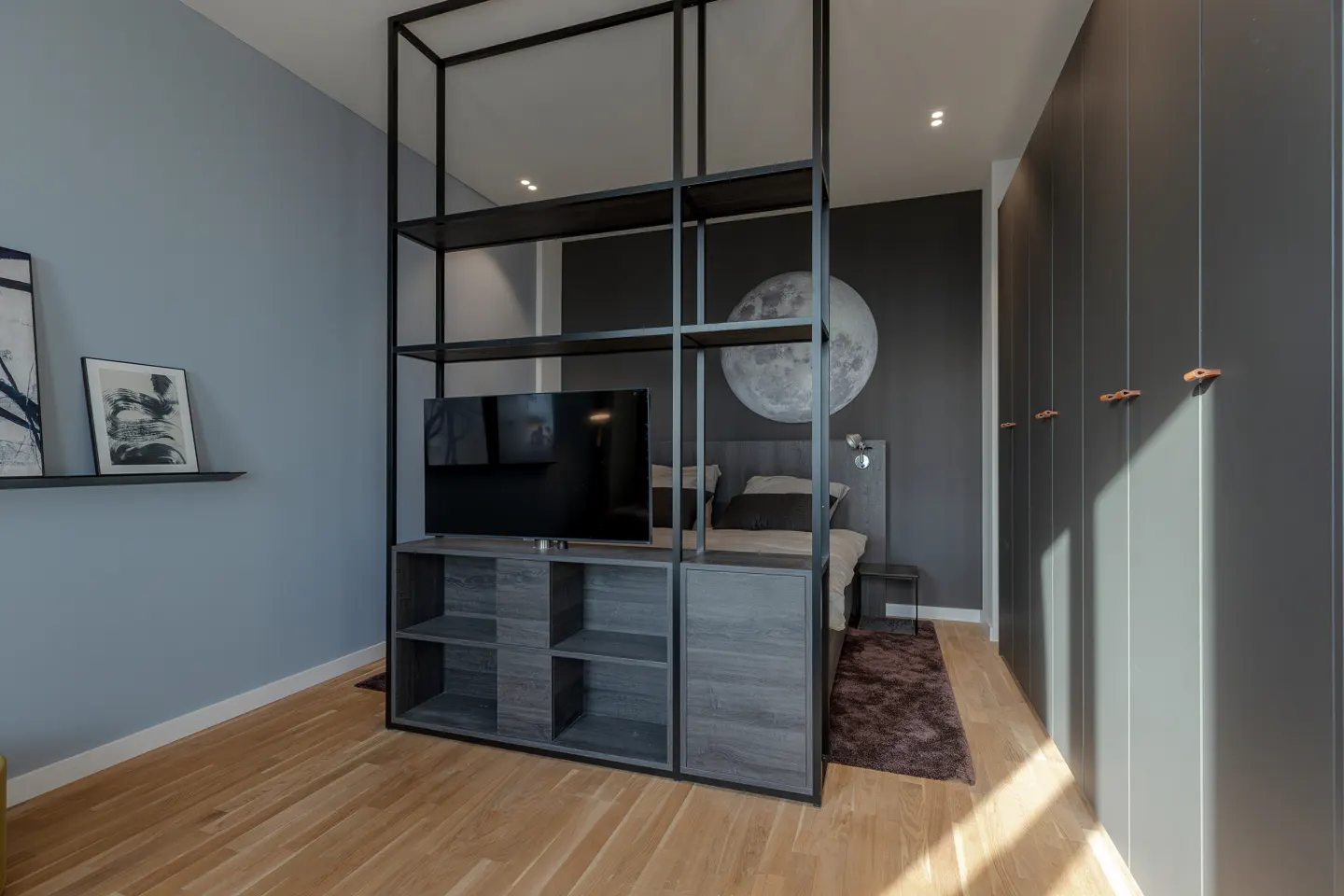 Bedroom with wood floors, blue and gray walls, and a black metal shelving unit with a TV. A bed with a moon art piece is behind the unit.