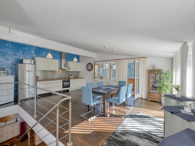 Bright, open-concept living space with a kitchen, dining table with blue chairs, and wood floors. A staircase with metal railings is visible.