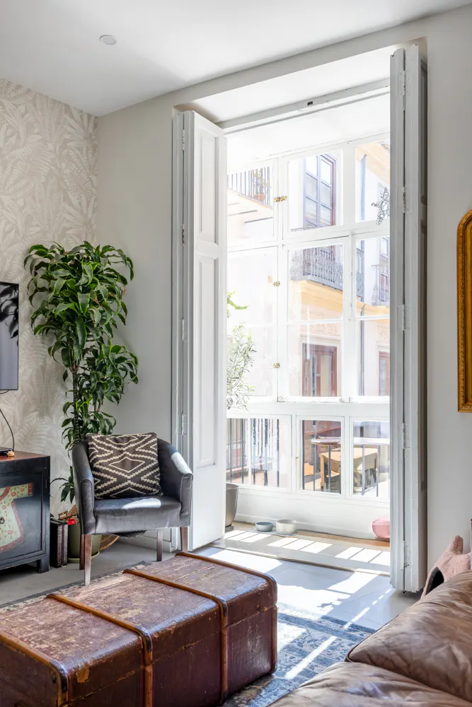 Living room with a brown leather ottoman, gray chair, and a large window with white shutters overlooking a courtyard.