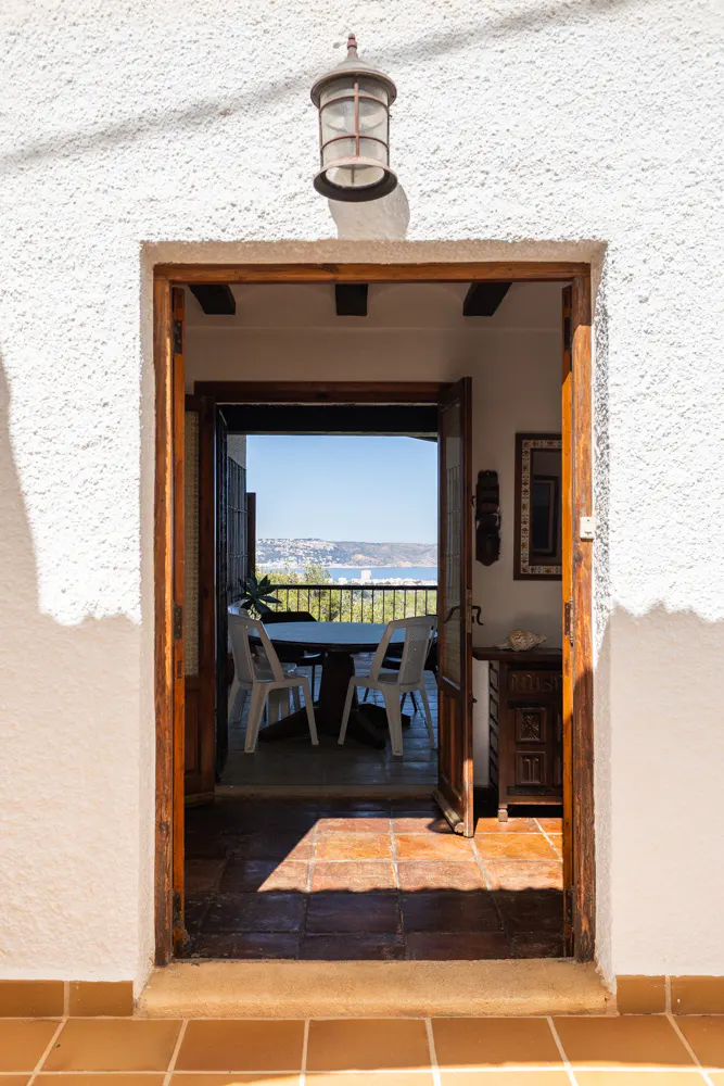 View through open wood doors to a patio with table, chairs, and ocean view. White stucco walls and tile floor.