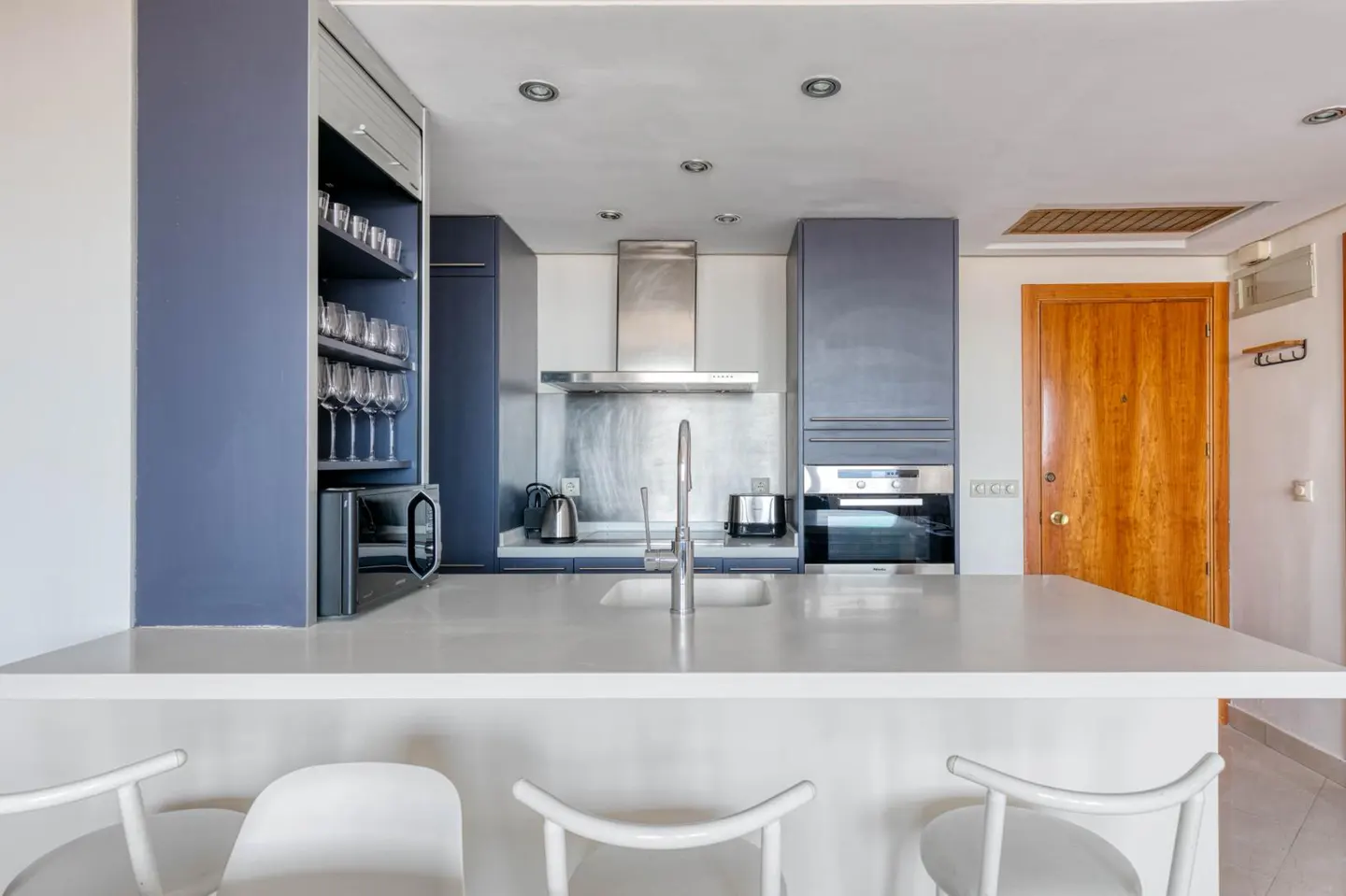 Modern kitchen with white countertop island and three white chairs. Blue cabinets, stainless steel appliances, and a wooden door are visible.