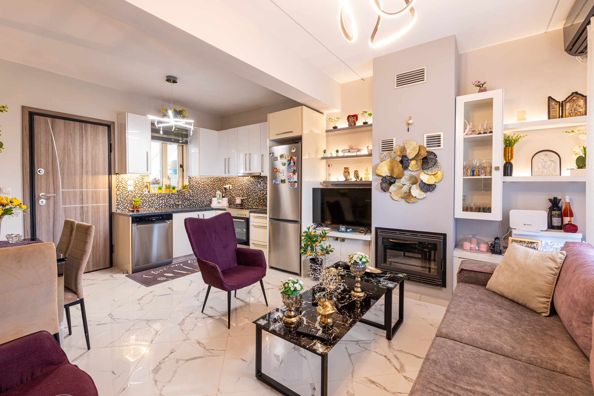 Modern open-plan living room with a purple armchair, marble coffee table, fireplace, and a white kitchen in the background. [AI-generated text]