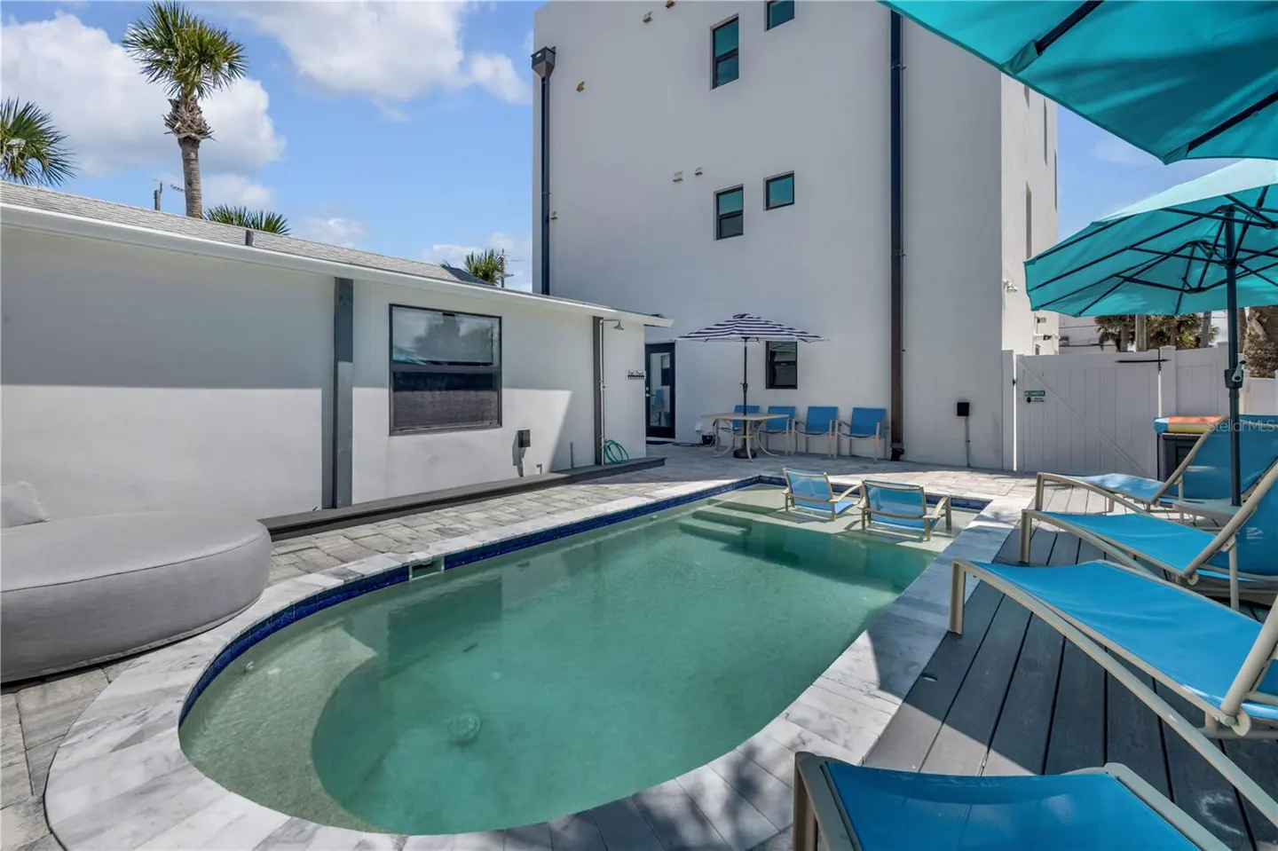 Outdoor pool area with blue lounge chairs, a table with chairs under a striped umbrella, and a white building in the background.