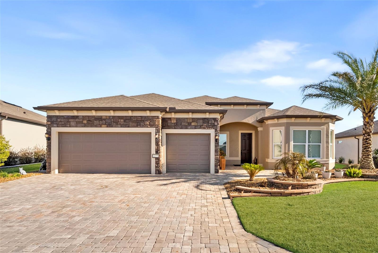 Beige single-story house with a brown roof, stone accents, two-car garage, and a brick driveway under a blue sky.