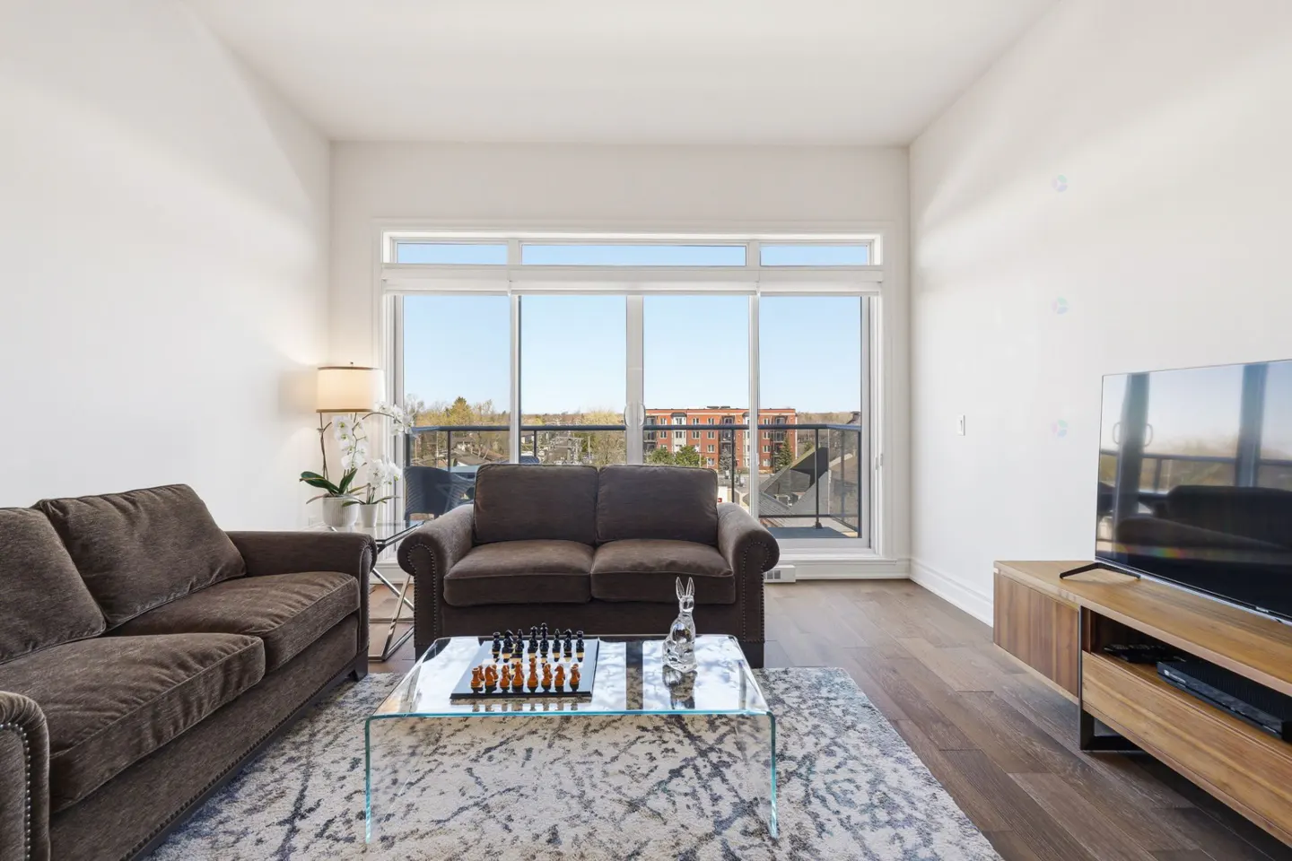 Bright living room with brown sofas, glass table with chess set, and large window with balcony view.