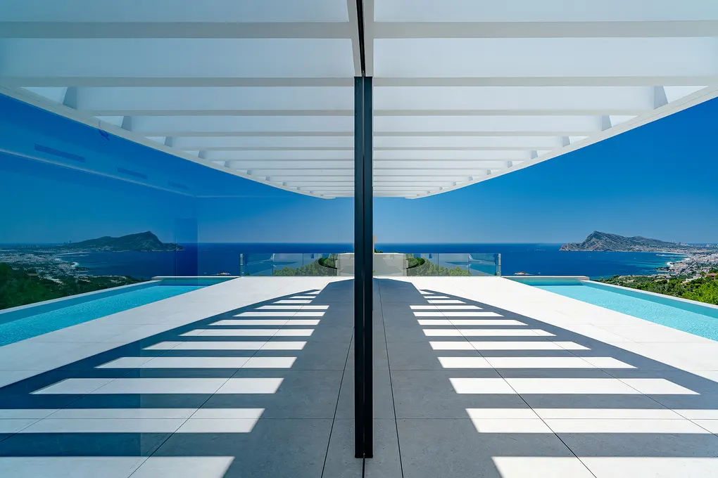 Modern home exterior with a pool, white pergola, and ocean view. Shadows create a striped pattern on the tiled patio.