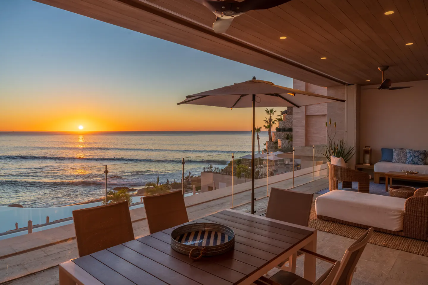 Ocean view from a covered patio with a wood table and chairs. A sunset is visible over the water.