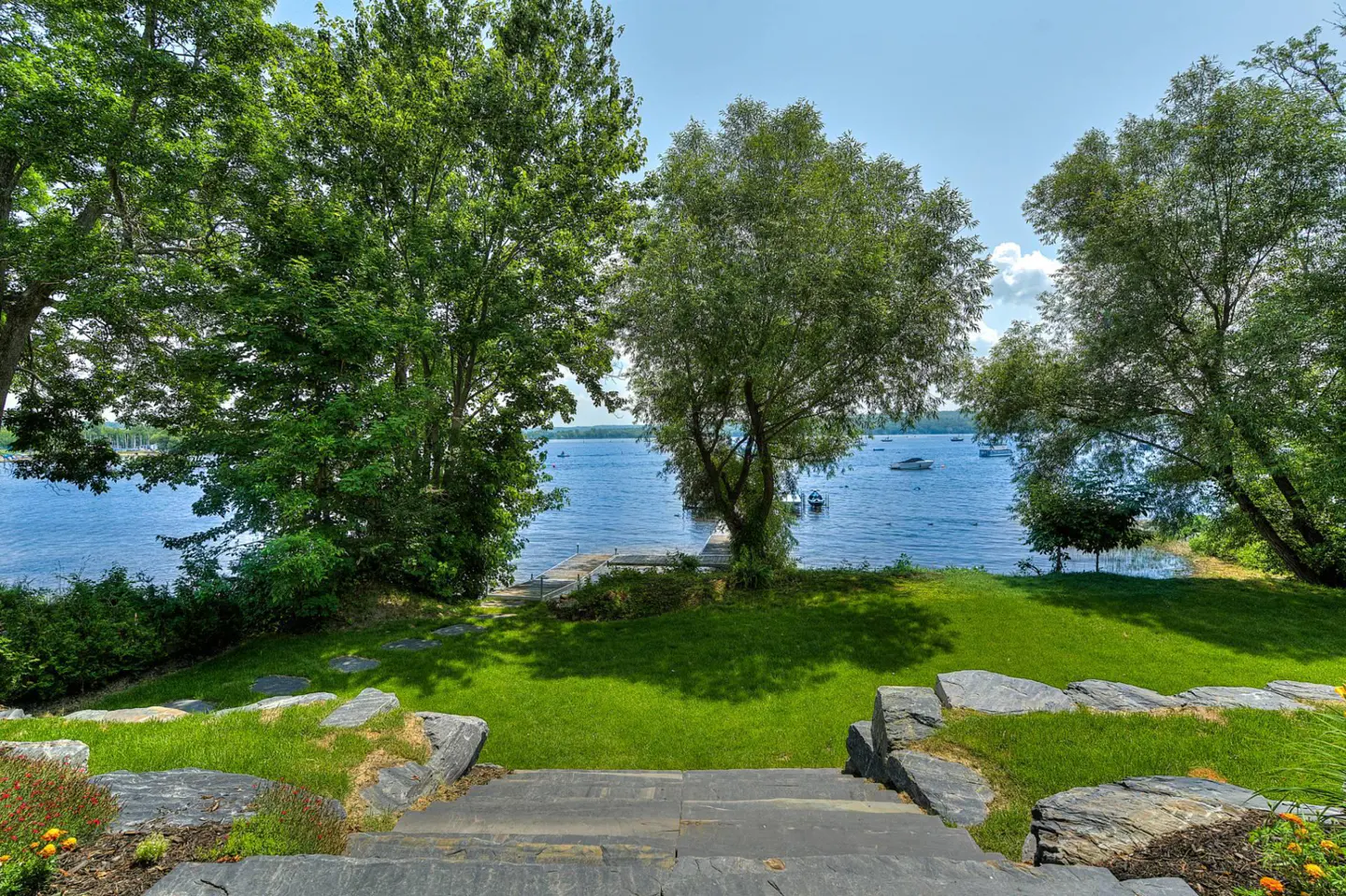 View from stone steps down to a green lawn, trees, and a blue lake with boats under a blue sky.