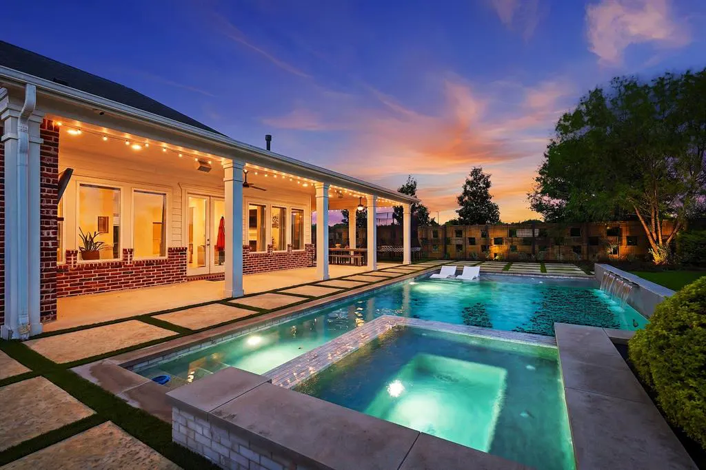 Outdoor pool and spa at dusk. White lounge chairs sit poolside. String lights illuminate the patio of a brick house.