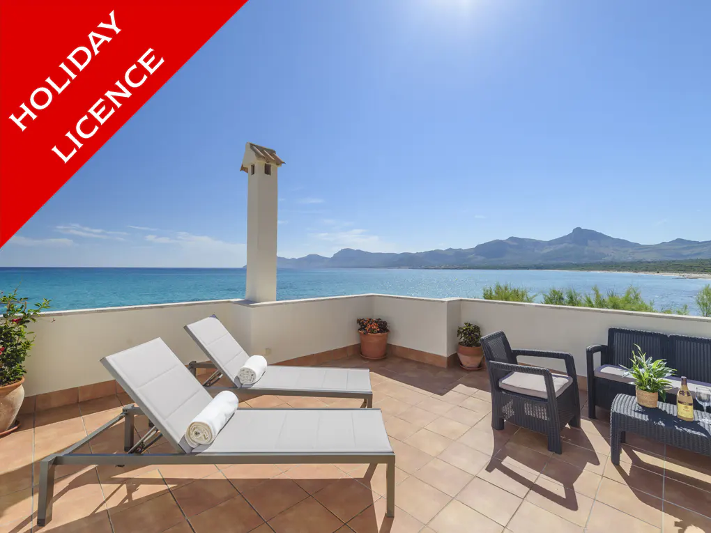 Terrace with two sunbeds, chairs, and a table overlooking the ocean and mountains under a blue sky. "Holiday Licence" banner.