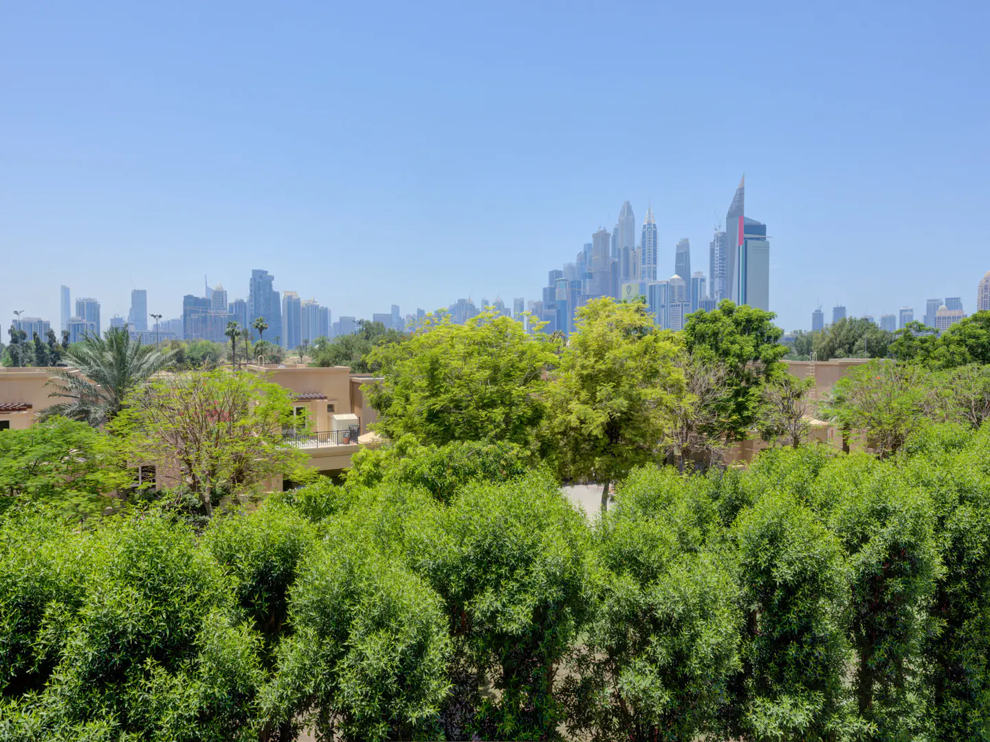 View of Dubai skyline over lush green trees and beige buildings under a clear blue sky.