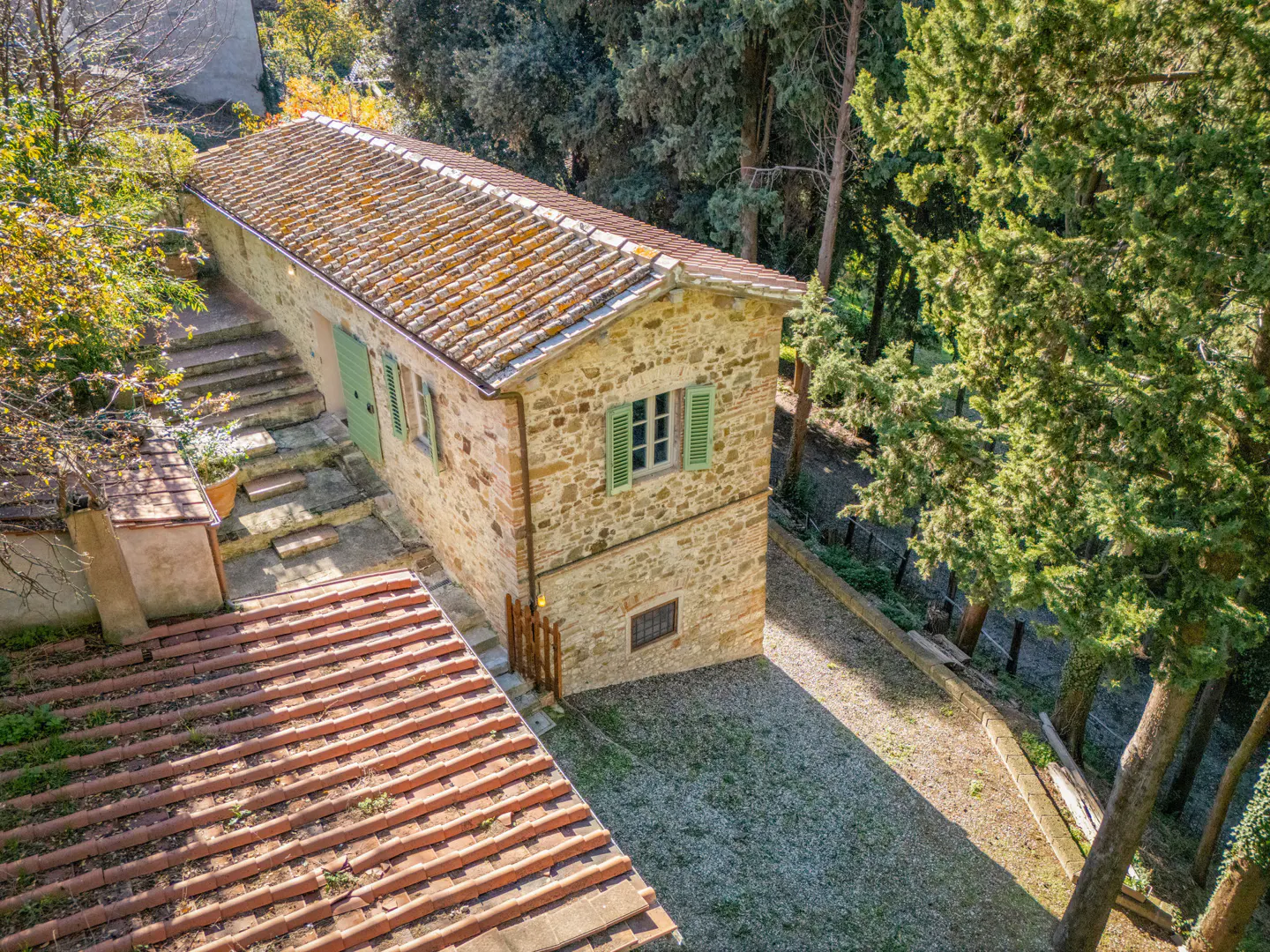 High-angle view of a rustic stone cottage with a terracotta tile roof, green shutters, and stone steps in a wooded area.