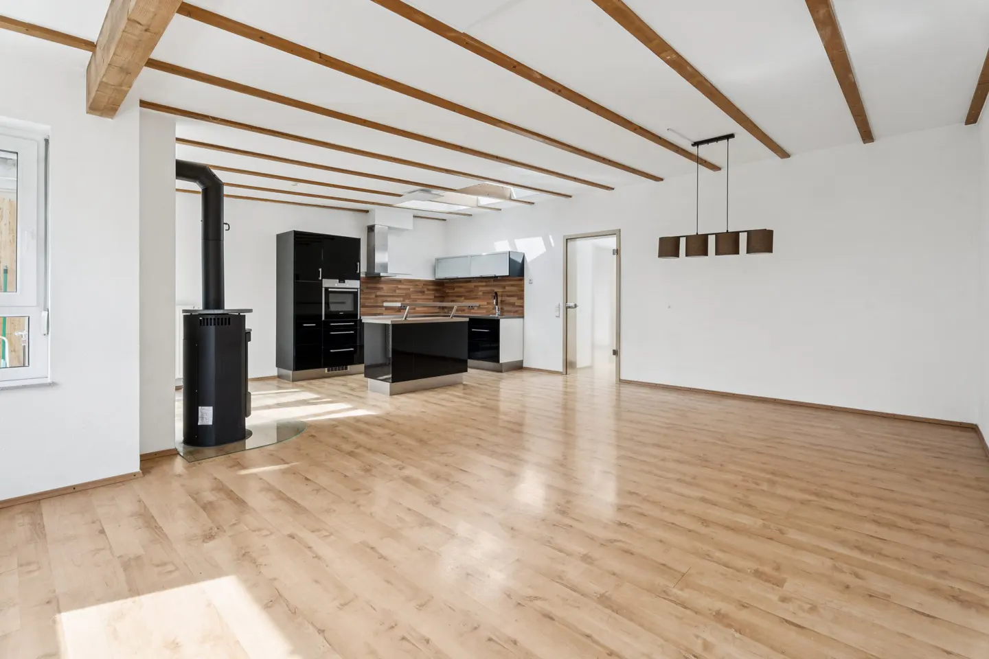 Bright, open-concept kitchen and living space with wood floors, white walls, and exposed wood beams. Black kitchen cabinets and wood-burning stove.