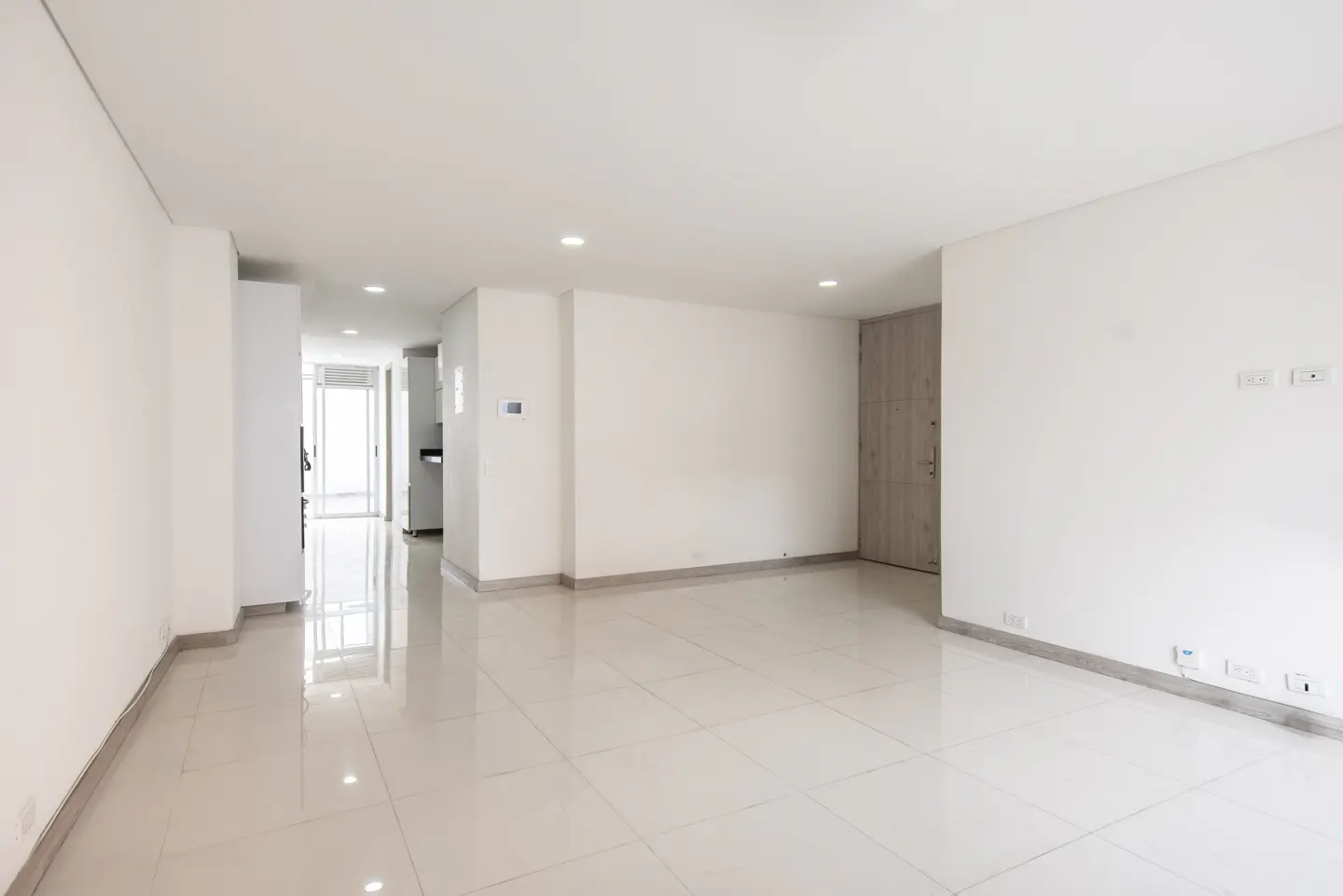 Bright, empty apartment with white walls and tile floors. A doorway leads to a kitchen with stainless steel appliances.