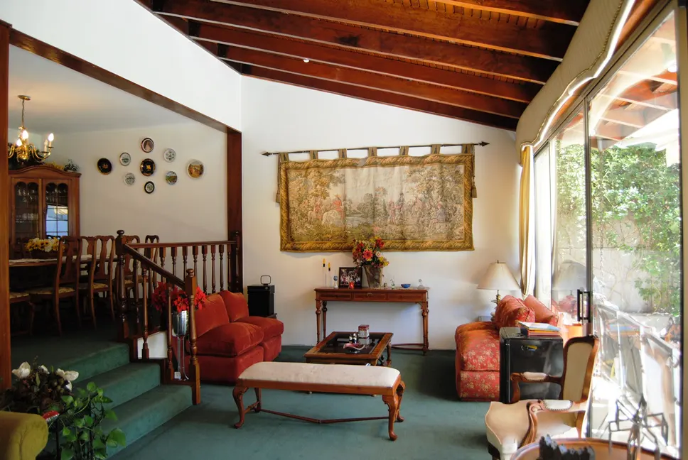A living room with a tapestry, red sofas, and a wooden ceiling. A dining room is visible in the background.