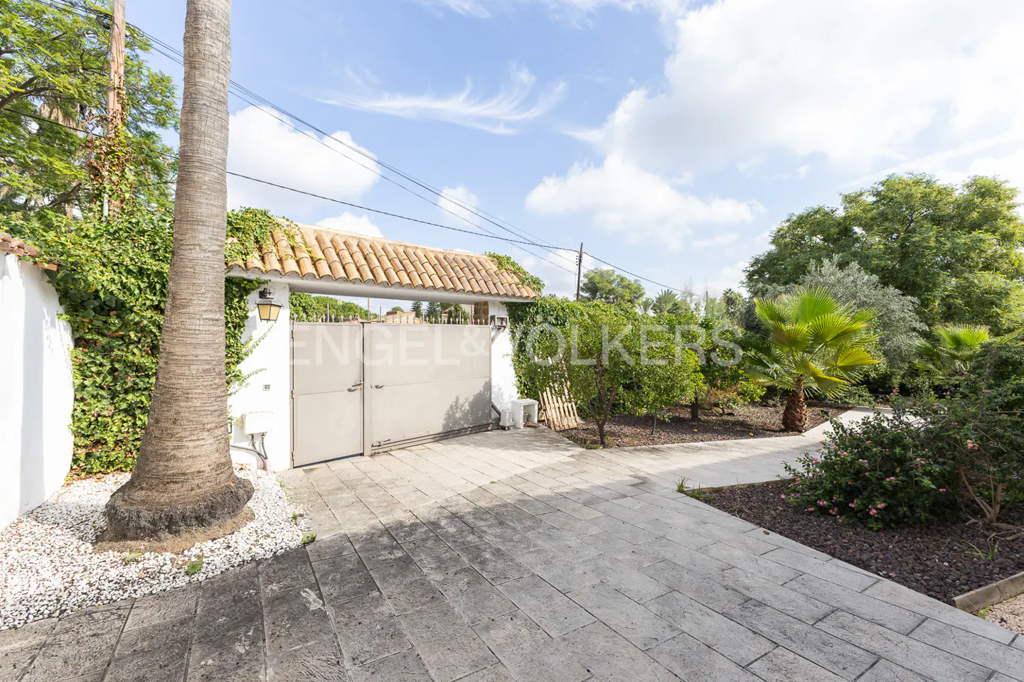 Gated driveway leading to a property with lush greenery and a blue sky backdrop.