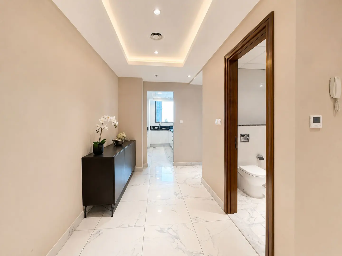 Hallway view with white marble floors, beige walls, and recessed lighting. A dark cabinet with flowers sits on the left, and a bathroom is visible through a doorway on the right.