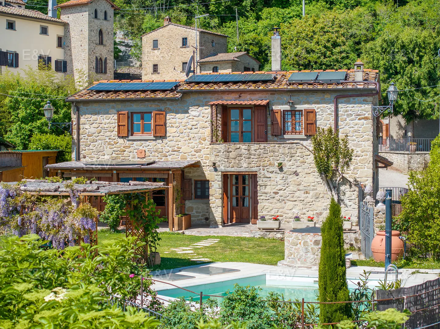 Stone house with brown shutters and solar panels, surrounded by greenery and a pool.