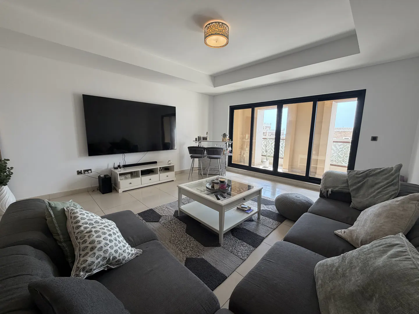 Living room with gray sectional sofa, white coffee table on a gray rug, and a large TV. Sliding glass doors lead to a balcony.