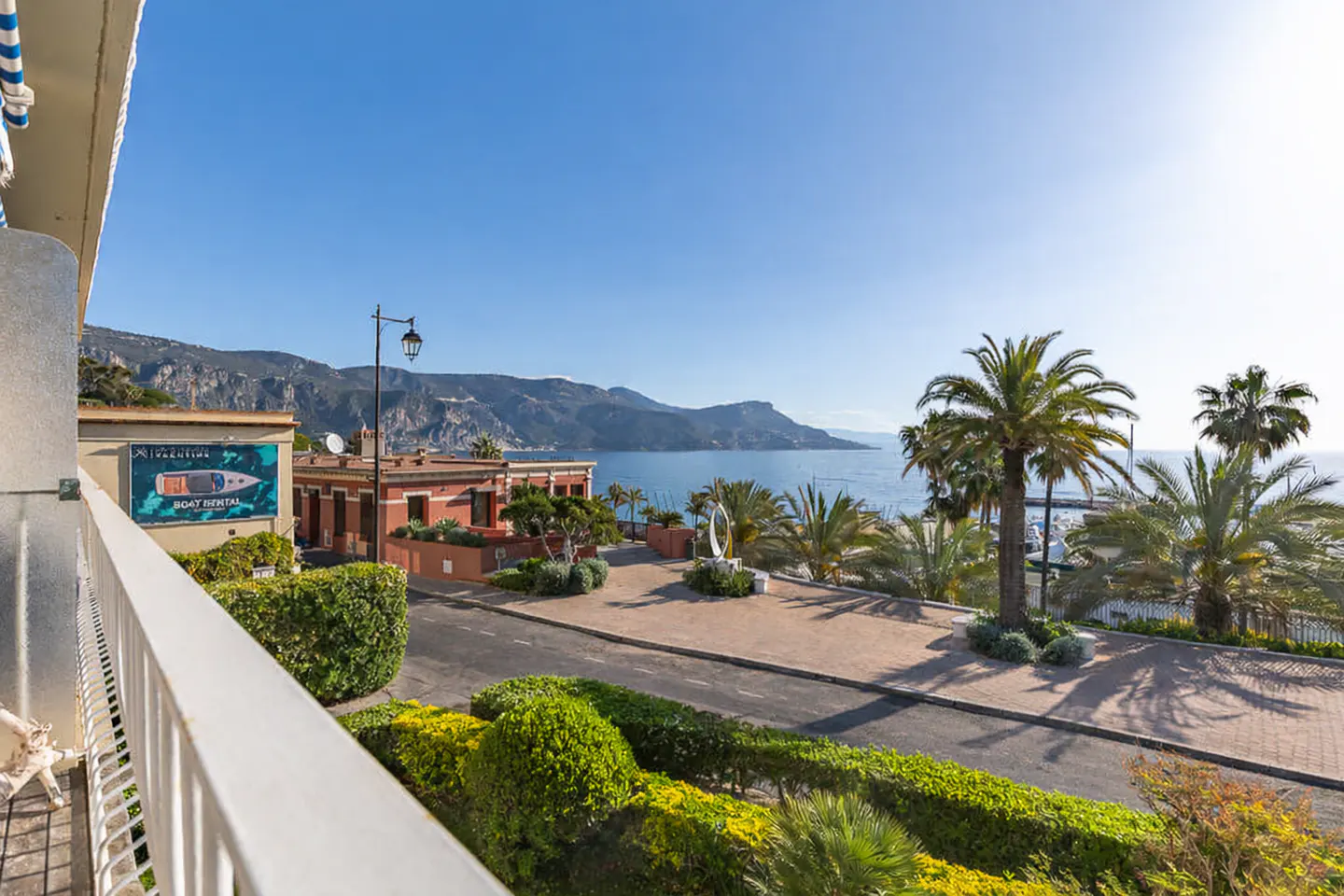 View from a balcony overlooking a coastal town with palm trees, mountains, and the sea under a clear blue sky. A "Boat Rental" sign is visible.