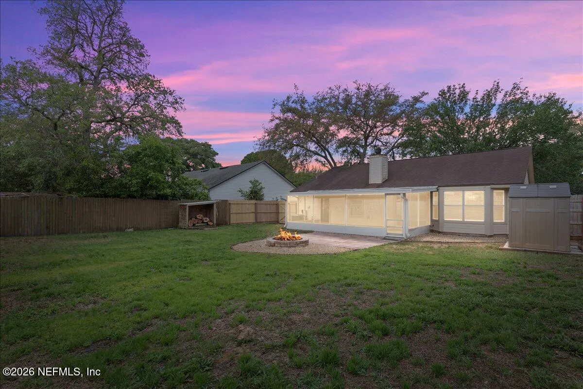 Backyard with green grass, wooden fence, fire pit with flames, and a screened-in porch at dusk with a pink and purple sky.