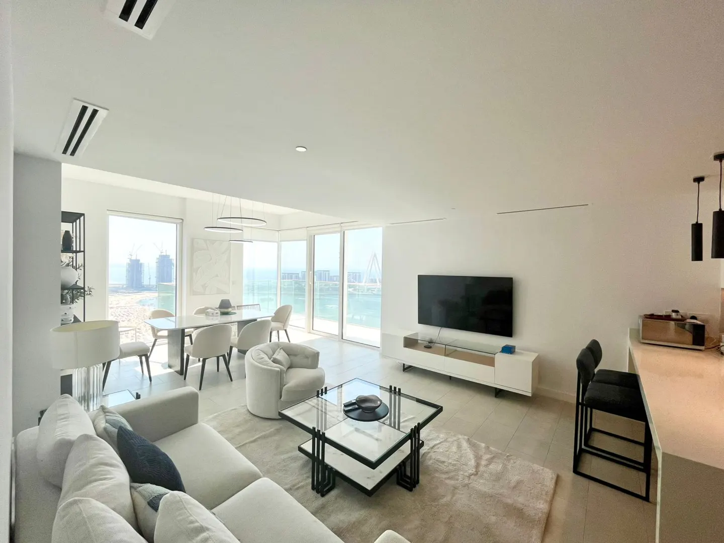 Bright, modern living room with white sofa, dining table, TV, and ocean view. Neutral tones create a clean, airy space.