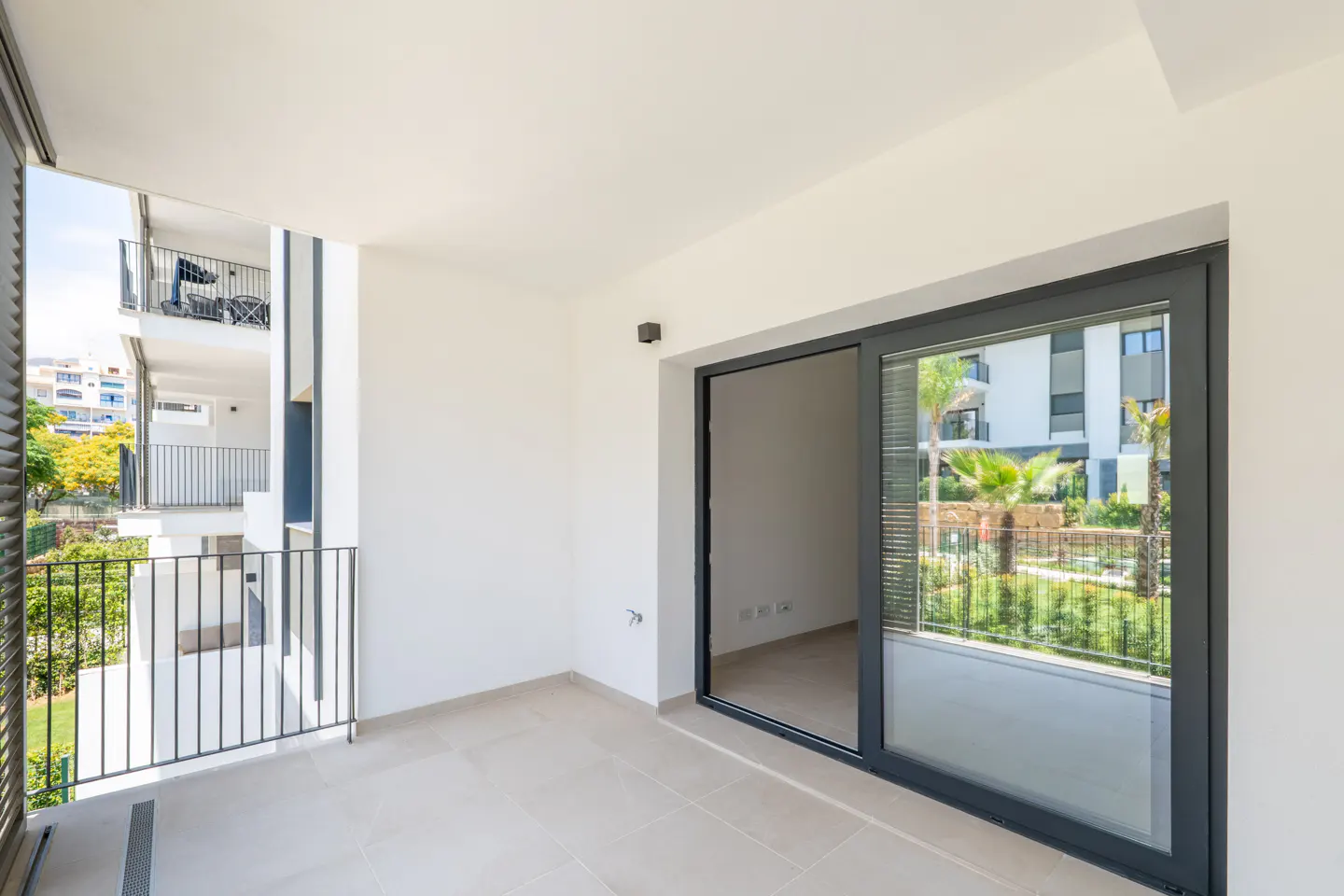 Modern apartment balcony with light tile flooring, white walls, and large sliding glass doors overlooking a garden area.