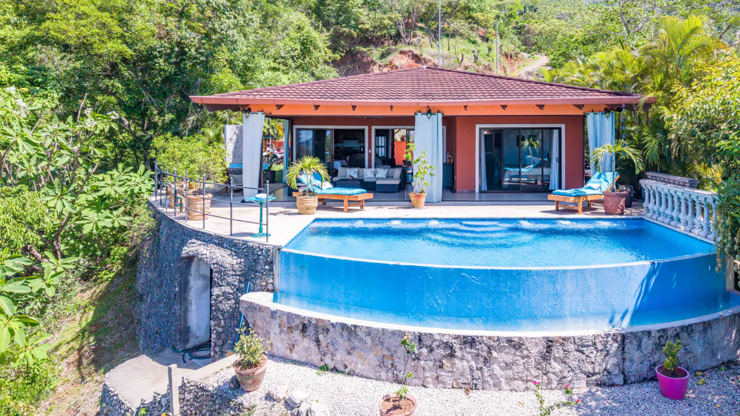 Exterior view of a terracotta house with a pool, lounge chairs, and lush green trees in the background.