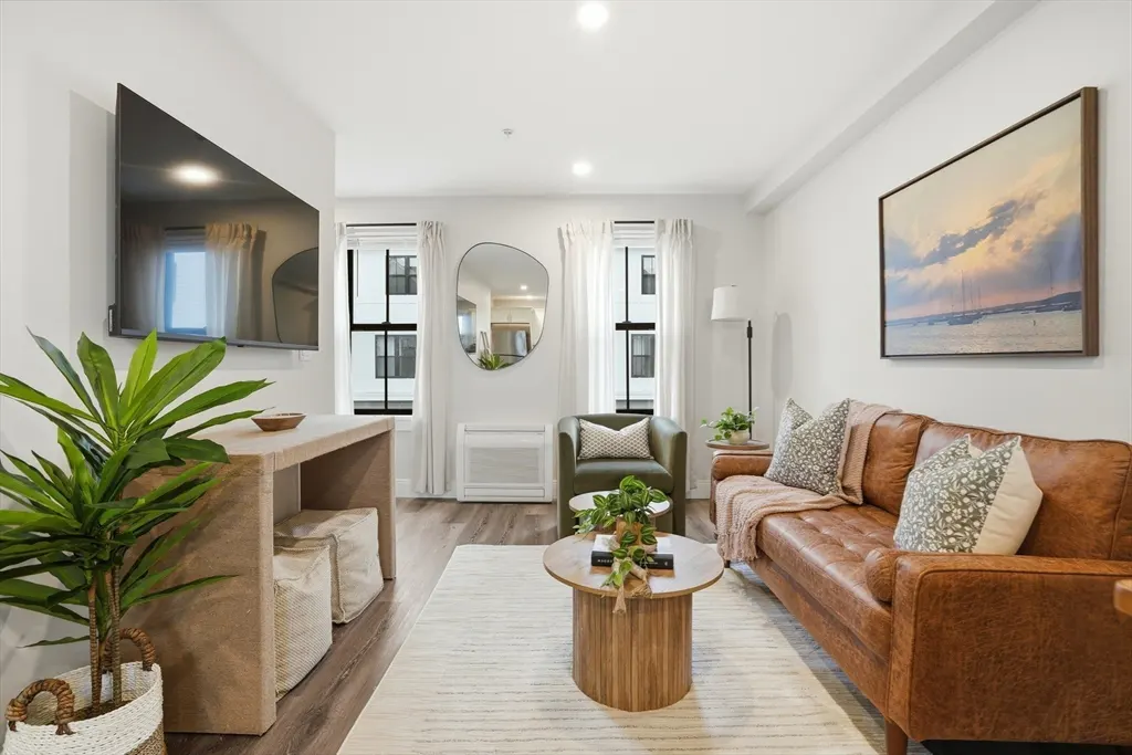 Bright living room with a brown leather sofa, green armchair, and a wooden coffee table on a striped rug. A large TV is mounted on the wall.