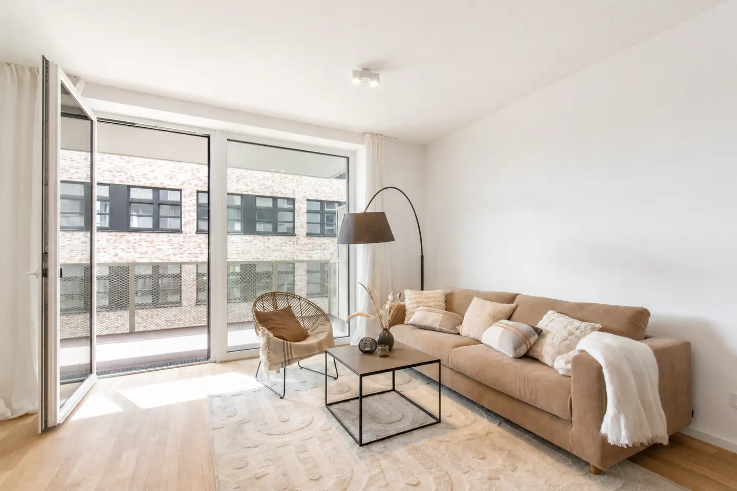 Modern living room with a beige sofa, rattan chair, coffee table, and floor lamp, featuring large windows to a balcony.