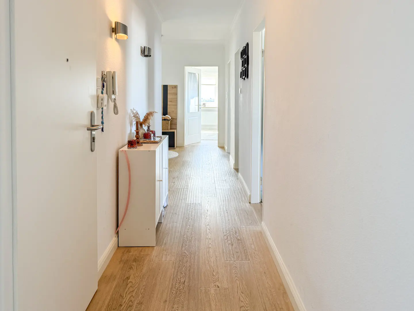 Bright hallway with light wood floors and white walls. A white cabinet sits near the entrance with keys hanging on the wall.