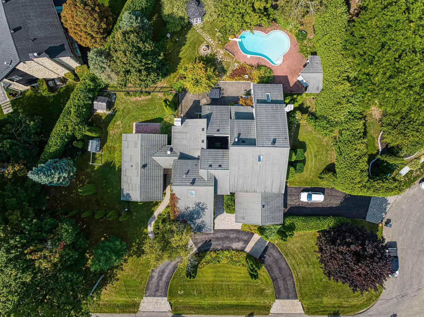 Aerial view of a large, modern house with a gray roof, a circular driveway, and a heart-shaped pool surrounded by lush greenery.