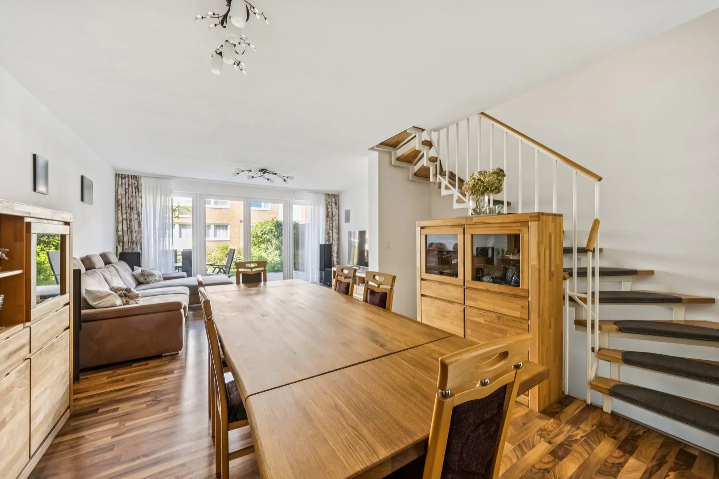 Bright, open-plan living and dining area with wood floors, a long wooden table, brown sofa, and staircase with white railings.