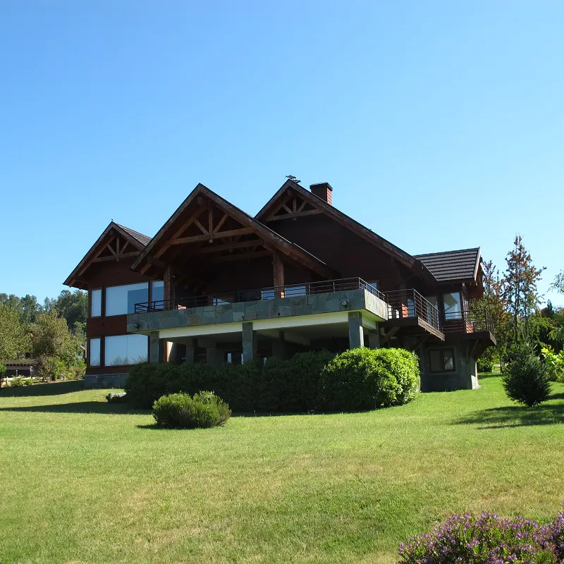 A large, brown, two-story house with a stone foundation and a green lawn under a clear blue sky.