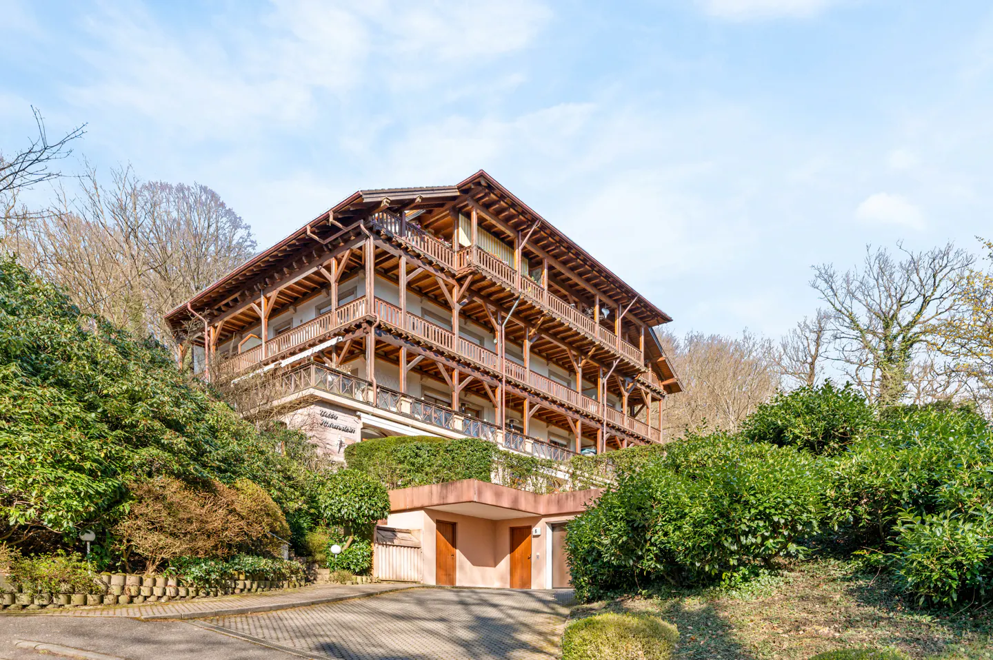 A three-story building with wooden balconies and a garage, surrounded by green bushes.