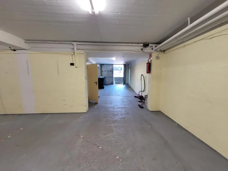 Empty underground garage with pale yellow walls, concrete floor, exposed pipes, and a doorway leading to an outdoor exit.