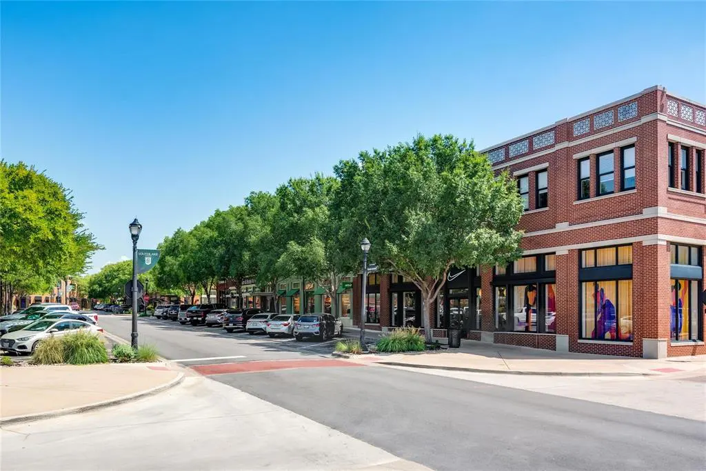 Street view of a commercial district with brick buildings, trees, parked cars, and a blue sky.