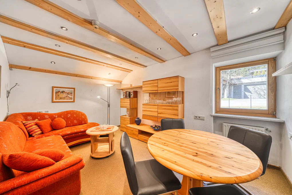 Living room with orange sofa, wooden table and chairs, and built-in wooden cabinets. The ceiling has wooden beams and recessed lighting.