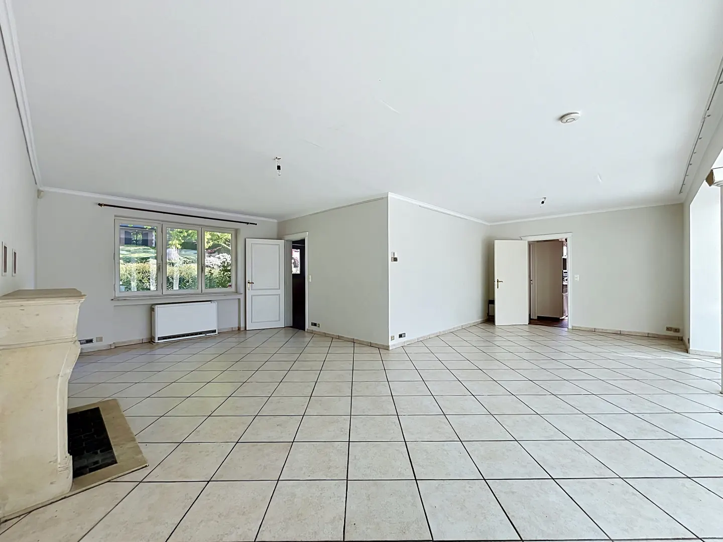Bright, empty living room with white walls, tile floor, and a fireplace. A window and two open doors are visible.