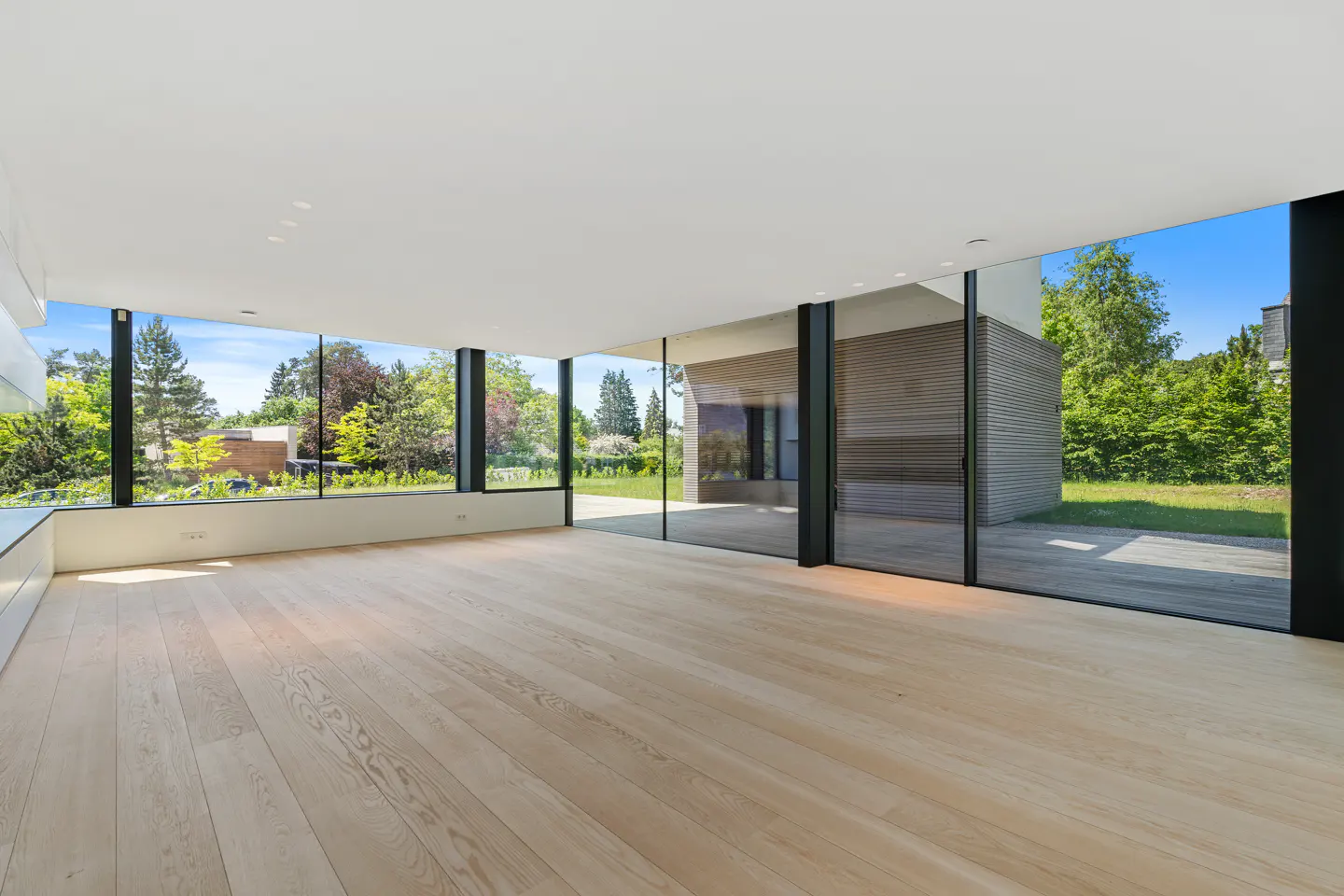 Empty modern living room with light wood floors, floor-to-ceiling glass walls, and a view of a lush green backyard.
