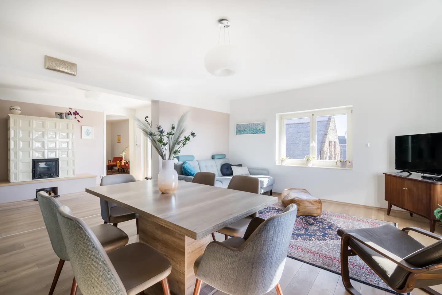 Bright living room with a dining table, chairs, sofa, fireplace, and TV. A vase of flowers sits on the table. Sunlight streams through the window.
