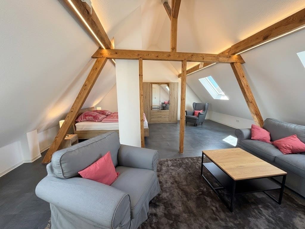 Attic apartment with exposed wooden beams, gray sofas with pink pillows, and a bedroom visible through a doorway.