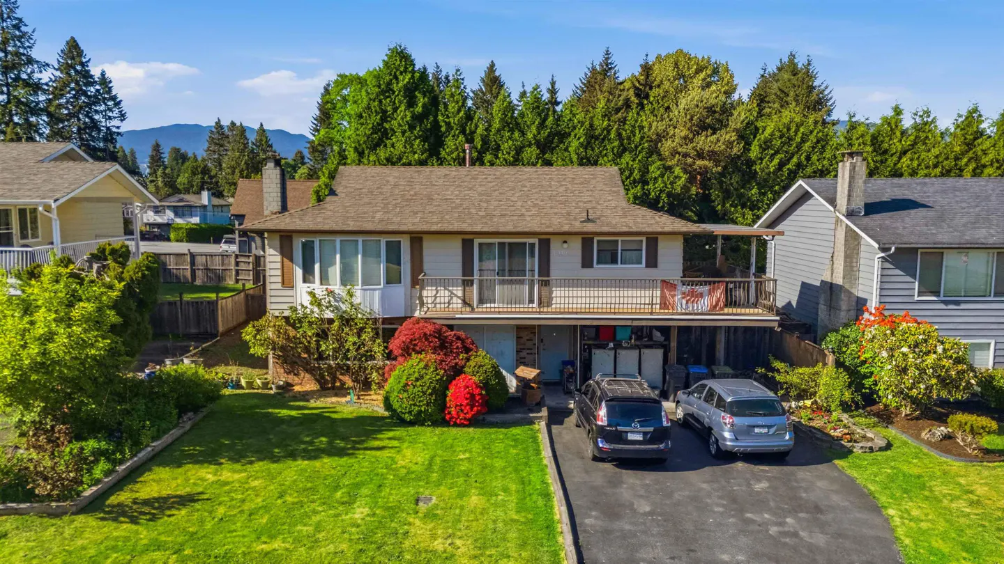 Two-story house with a brown roof, beige siding, and a balcony with a Canadian flag. Two cars are parked in the driveway.